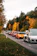 A lineup of luxury sports cars is parked along a narrow road. The cars are in various colors, including white, orange, red, and black, and are positioned in a single file. The scene is set during autumn with vibrant foliage, as the trees along the road display a mix of green and golden-yellow leaves.