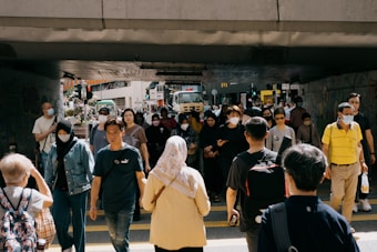 A bustling urban street scene under an overpass with a diverse group of people walking in different directions. Many individuals are wearing face masks, suggesting awareness of health measures. The background displays city infrastructure, including vehicles and buildings with some greenery on the side.