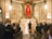A wedding ceremony taking place inside a church, with a bride and groom standing before the altar as a priest officiates. The bride is dressed in a white gown with a veil while the groom is in a black suit. The church is adorned with religious artwork, including a prominent crucifix, and filled with guests seated in pews. The interior features high arches, detailed frescoes, and a warm lighting ambiance.