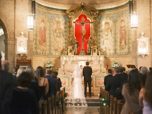 A wedding ceremony taking place inside a church, with a bride and groom standing before the altar as a priest officiates. The bride is dressed in a white gown with a veil while the groom is in a black suit. The church is adorned with religious artwork, including a prominent crucifix, and filled with guests seated in pews. The interior features high arches, detailed frescoes, and a warm lighting ambiance.