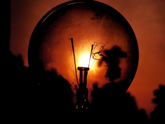 A close-up of a light bulb against a warm, orange background. The bulb appears dark and silhouetted, with intricate details of the filament visible inside. The background gives a fiery impression, enhancing the contrast with the bulb.