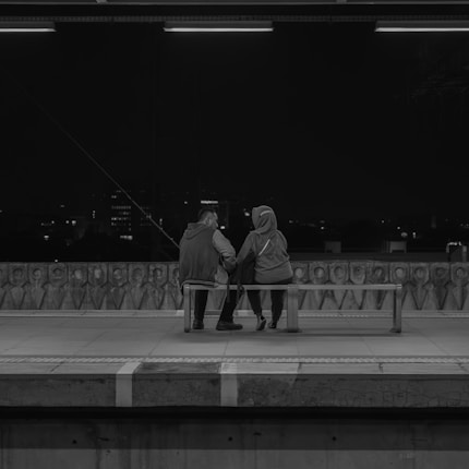 Two people are sitting together on a bench at a train platform during nighttime. They are facing away, looking out at the distant cityscape, which is dimly lit in the background. The setting is calm and intimate, emphasizing a sense of togetherness.