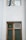 A section of a building facade featuring two windows. The upper window has a wooden frame and is reflecting tree branches. The lower window, also with a wooden frame, includes a decorative green glass pane and a small louvered vent on the side.