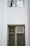 A section of a building facade featuring two windows. The upper window has a wooden frame and is reflecting tree branches. The lower window, also with a wooden frame, includes a decorative green glass pane and a small louvered vent on the side.