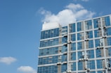 A modern building with a glass facade reflecting the clear blue sky. The structure is fitted with several air conditioning units visible on the exterior walls.