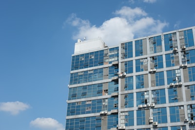 A modern building with a glass facade reflecting the clear blue sky. The structure is fitted with several air conditioning units visible on the exterior walls.