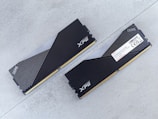 Two DDR5 RAM modules with black and silver heatsinks are positioned on a flat, light gray surface. One module is viewed from the top showing the XPG logo and a pattern of diagonal lines, while the other displays a label with technical specifications.