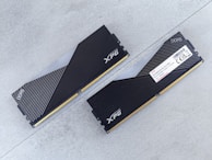 Two DDR5 RAM modules with black and silver heatsinks are positioned on a flat, light gray surface. One module is viewed from the top showing the XPG logo and a pattern of diagonal lines, while the other displays a label with technical specifications.