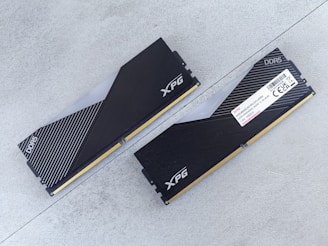 Two DDR5 RAM modules with black and silver heatsinks are positioned on a flat, light gray surface. One module is viewed from the top showing the XPG logo and a pattern of diagonal lines, while the other displays a label with technical specifications.