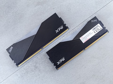 Two DDR5 RAM modules with black and silver heatsinks are positioned on a flat, light gray surface. One module is viewed from the top showing the XPG logo and a pattern of diagonal lines, while the other displays a label with technical specifications.