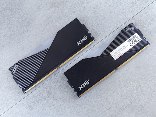 Two DDR5 RAM modules with black and silver heatsinks are positioned on a flat, light gray surface. One module is viewed from the top showing the XPG logo and a pattern of diagonal lines, while the other displays a label with technical specifications.