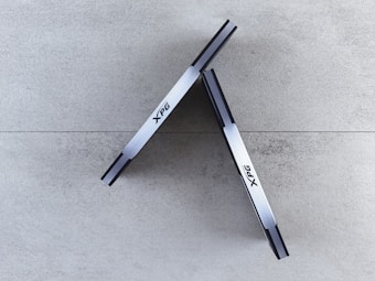 Two sleek, metallic sticks labeled with 'XPG' are positioned against a neutral, textured background forming an inverted 'V' shape at the top. The background consists of large stone-like tiles.
