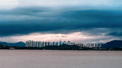 A cityscape with a high-rise apartment buildings lined up along the shoreline. The skyline is set against a backdrop of dark, brooding clouds and distant hills. There is a large body of water in the foreground, possibly a lake or river.