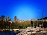 A person is fishing on a rocky embankment near a body of water, with a backdrop of a cityscape featuring modern buildings. The sky is clear and blue, and a group of people with bicycles is nearby, suggesting a leisurely, outdoor setting.