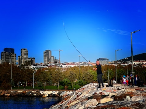 A person is fishing on a rocky embankment near a body of water, with a backdrop of a cityscape featuring modern buildings. The sky is clear and blue, and a group of people with bicycles is nearby, suggesting a leisurely, outdoor setting.