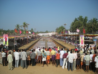 A large crowd of people gather around a long, narrow water-filled track, possibly for a traditional bull racing event. Colorful flags and banners decorate the area, with palm trees and other greenery in the background. Spectators line both sides of the track, some sitting and others standing, observing the event.