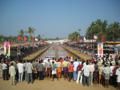 A large crowd of people gather around a long, narrow water-filled track, possibly for a traditional bull racing event. Colorful flags and banners decorate the area, with palm trees and other greenery in the background. Spectators line both sides of the track, some sitting and others standing, observing the event.