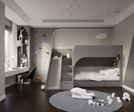 A modern children's bedroom featuring a sleek gray bunk bed with a built-in slide. The room includes a climbing wall with colorful holds and a study area with a desk by the window. Decor includes white toys, a playful whale figure on a textured gray carpet, and several decorative elements on shelves. Soft lighting and a minimalist design create a warm and inviting atmosphere.