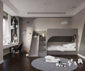 A modern children's bedroom featuring a sleek gray bunk bed with a built-in slide. The room includes a climbing wall with colorful holds and a study area with a desk by the window. Decor includes white toys, a playful whale figure on a textured gray carpet, and several decorative elements on shelves. Soft lighting and a minimalist design create a warm and inviting atmosphere.