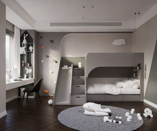 A modern children's bedroom featuring a sleek gray bunk bed with a built-in slide. The room includes a climbing wall with colorful holds and a study area with a desk by the window. Decor includes white toys, a playful whale figure on a textured gray carpet, and several decorative elements on shelves. Soft lighting and a minimalist design create a warm and inviting atmosphere.
