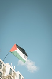 A flag with red, black, white, and green colors is waving from a flagpole attached to a building. The sky is clear and blue, dominated by a single small cloud in the background. The architecture of the building shows modern, rectangular windows and a pale facade.