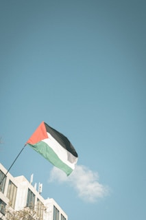 A flag with red, black, white, and green colors is waving from a flagpole attached to a building. The sky is clear and blue, dominated by a single small cloud in the background. The architecture of the building shows modern, rectangular windows and a pale facade.
