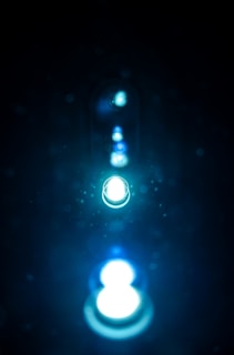 A series of glowing lights appear in a line, creating a sense of depth. The lights are surrounded by an ethereal glow with particles reflecting the light, set against a dark background.