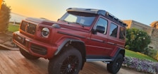 A red luxury SUV is parked on a wooden platform surrounded by greenery and flowers. The vehicle has a rugged design with large tires and a roof rack. In the background, there is a glimpse of the ocean and a stone building.