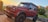 A red luxury SUV is parked on a wooden platform surrounded by greenery and flowers. The vehicle has a rugged design with large tires and a roof rack. In the background, there is a glimpse of the ocean and a stone building.