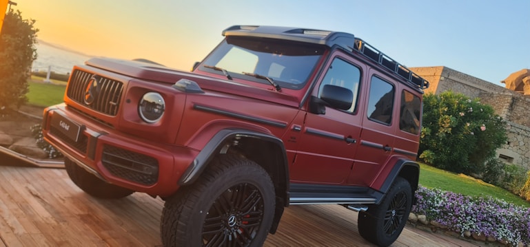 A red luxury SUV is parked on a wooden platform surrounded by greenery and flowers. The vehicle has a rugged design with large tires and a roof rack. In the background, there is a glimpse of the ocean and a stone building.