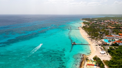 Aerial view of a tropical beach with turquoise waters extending towards the horizon. A small boat creates a wake as it travels through the clear sea. The shoreline is lined with white sandy beaches and resorts surrounded by lush greenery. A long pier juts into the water, and the area is dotted with palm trees and residential buildings.