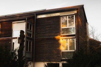 A wooden house with large windows reflecting the warm light of sunset. The exterior features horizontal wooden paneling, and there is a birdhouse mounted on a tree stump in the foreground. Surrounding the house are some trees and bushes, contributing to a natural setting.
