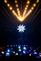 A captivating nighttime scene featuring a large, illuminated star-like structure suspended above, with strings of warm lights radiating upwards. Below, there are various colorful light installations, including patterns and abstract shapes, creating a vibrant visual spectacle.