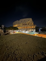 A nighttime view of the Colosseum in Rome, Italy, illuminated with warm lighting against a dark sky. The ancient structure stands prominently with its iconic arches, surrounded by a few trees and visible construction barriers in the foreground. There are a few people visible, indicating a lightly bustling atmosphere around the site.