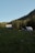 Wooden cabins with metal roofs are nestled in a lush green meadow surrounded by dense forest. A gentle slope leads up to the structures, and the scene is set against a clear blue sky. The sunlight softly illuminates parts of the forest and cabins, creating a tranquil atmosphere.