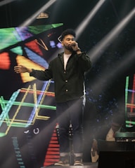 A man is singing on stage, holding a microphone in one hand and extending the other arm outward. He is dressed in a dark green shirt and jeans. Bright stage lights beam down on him, and a colorful geometric digital background is displayed behind. Another individual is partially visible in the background, playing a keyboard or similar instrument.