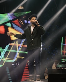 A man is singing on stage, holding a microphone in one hand and extending the other arm outward. He is dressed in a dark green shirt and jeans. Bright stage lights beam down on him, and a colorful geometric digital background is displayed behind. Another individual is partially visible in the background, playing a keyboard or similar instrument.