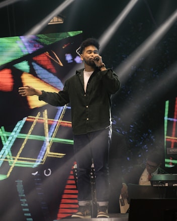 A man is singing on stage, holding a microphone in one hand and extending the other arm outward. He is dressed in a dark green shirt and jeans. Bright stage lights beam down on him, and a colorful geometric digital background is displayed behind. Another individual is partially visible in the background, playing a keyboard or similar instrument.