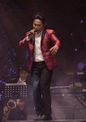 A singer wearing a red suit jacket and holding a microphone is performing on stage. In the background, a musician playing a saxophone is seated with a music stand in front of him. There is a keyboard and other musical equipment visible, and a smoky atmosphere enhances the stage lighting.