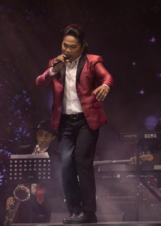 A singer wearing a red suit jacket and holding a microphone is performing on stage. In the background, a musician playing a saxophone is seated with a music stand in front of him. There is a keyboard and other musical equipment visible, and a smoky atmosphere enhances the stage lighting.