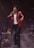 A singer wearing a red suit jacket and holding a microphone is performing on stage. In the background, a musician playing a saxophone is seated with a music stand in front of him. There is a keyboard and other musical equipment visible, and a smoky atmosphere enhances the stage lighting.