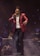 A singer wearing a red suit jacket and holding a microphone is performing on stage. In the background, a musician playing a saxophone is seated with a music stand in front of him. There is a keyboard and other musical equipment visible, and a smoky atmosphere enhances the stage lighting.