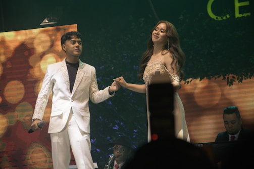 A man and a woman in formal attire are performing on stage. The man, wearing a white suit, holds a microphone, while the woman in a glamorous dress poses beside him. In the background, a musician plays the keyboard. The scene is set with warm lighting and decorative plants. The atmosphere appears celebratory and elegant.