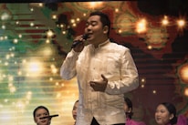 A man dressed in a traditional, cream-colored embroidered shirt is singing into a microphone on stage. Behind him, a colorful, abstract background features bright, star-like patterns. Several women in matching pink dresses are seated behind the singer, some with microphones, suggesting a choir or musical performance.