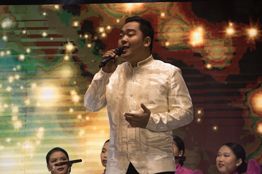A man dressed in a traditional, cream-colored embroidered shirt is singing into a microphone on stage. Behind him, a colorful, abstract background features bright, star-like patterns. Several women in matching pink dresses are seated behind the singer, some with microphones, suggesting a choir or musical performance.