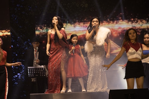 Several performers are on stage in a lively concert setting. Two women at the forefront are singing into microphones, dressed in elegant evening gowns, one in red and the other in silver. A young girl in a red dress stands between them, also holding a microphone. Other performers surround them, including a saxophonist in the background, wearing a hat and glasses. The stage is lit up, and the backdrop displays vivid colors and abstract designs.
