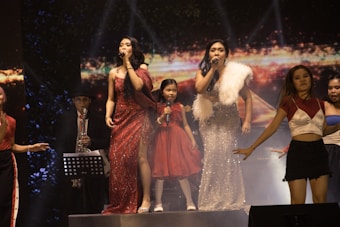 Several performers are on stage in a lively concert setting. Two women at the forefront are singing into microphones, dressed in elegant evening gowns, one in red and the other in silver. A young girl in a red dress stands between them, also holding a microphone. Other performers surround them, including a saxophonist in the background, wearing a hat and glasses. The stage is lit up, and the backdrop displays vivid colors and abstract designs.