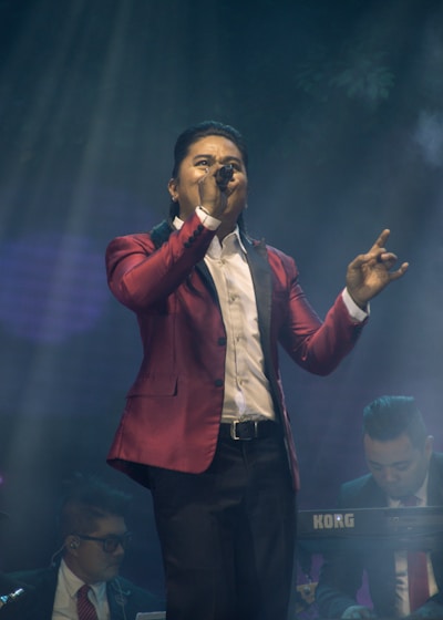 A performer in a red blazer is singing passionately into a microphone on stage. Two musicians are in the background, one playing a keyboard. The stage lighting creates an atmospheric effect with beams of light and a smoky ambiance.
