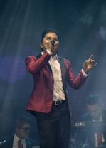 A performer in a red blazer is singing passionately into a microphone on stage. Two musicians are in the background, one playing a keyboard. The stage lighting creates an atmospheric effect with beams of light and a smoky ambiance.