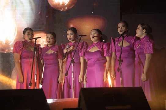 A group of six women stand in elegant magenta dresses with intricate lace details, performing on stage with microphones. They appear to be engaged in a musical performance, possibly singing together.